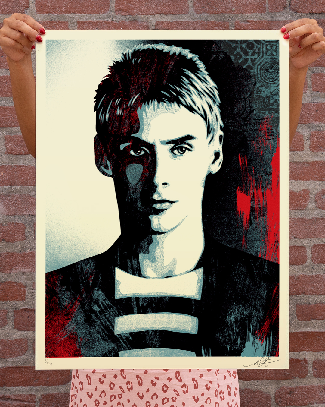 Hands holding a pop art print of a young man with short dark hair, in black, white, and red tones, against a brick wall.
