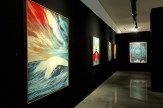 A modern art gallery with a black wall displaying four framed art prints. The first is a large, colorful abstract landscape…
