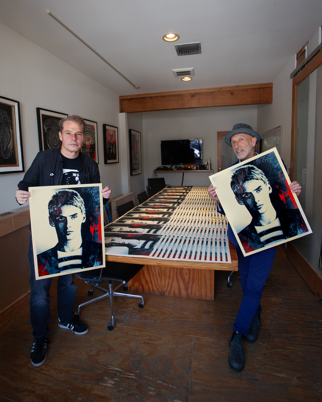 Two men holding pop art prints of a young man's portrait, standing in a room with a long table covered in similar prints.