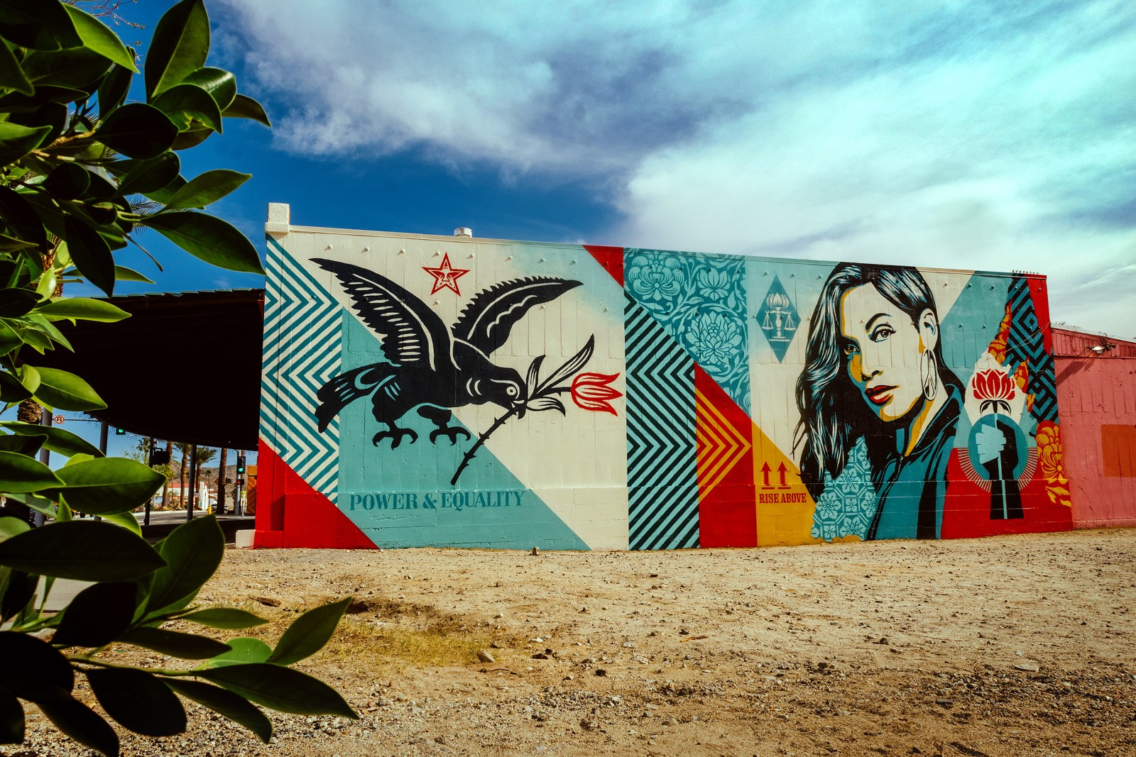 A colorful mural on a building featuring a bird, a woman's portrait, and various patterns in red, blue, yellow, and cream…