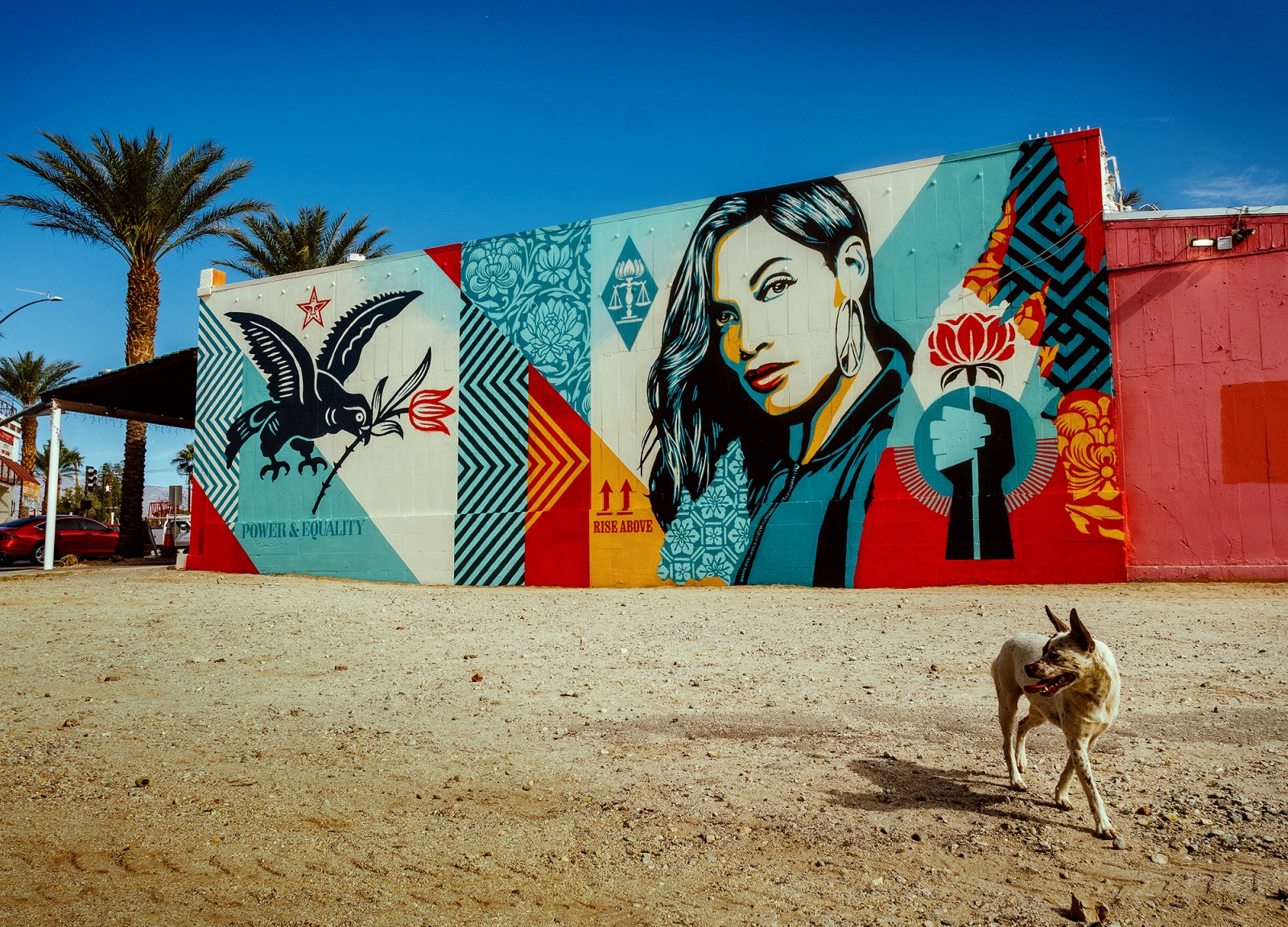 A colorful mural on a building featuring a bird, a woman's portrait, and various patterns in red, blue, yellow, and cream…