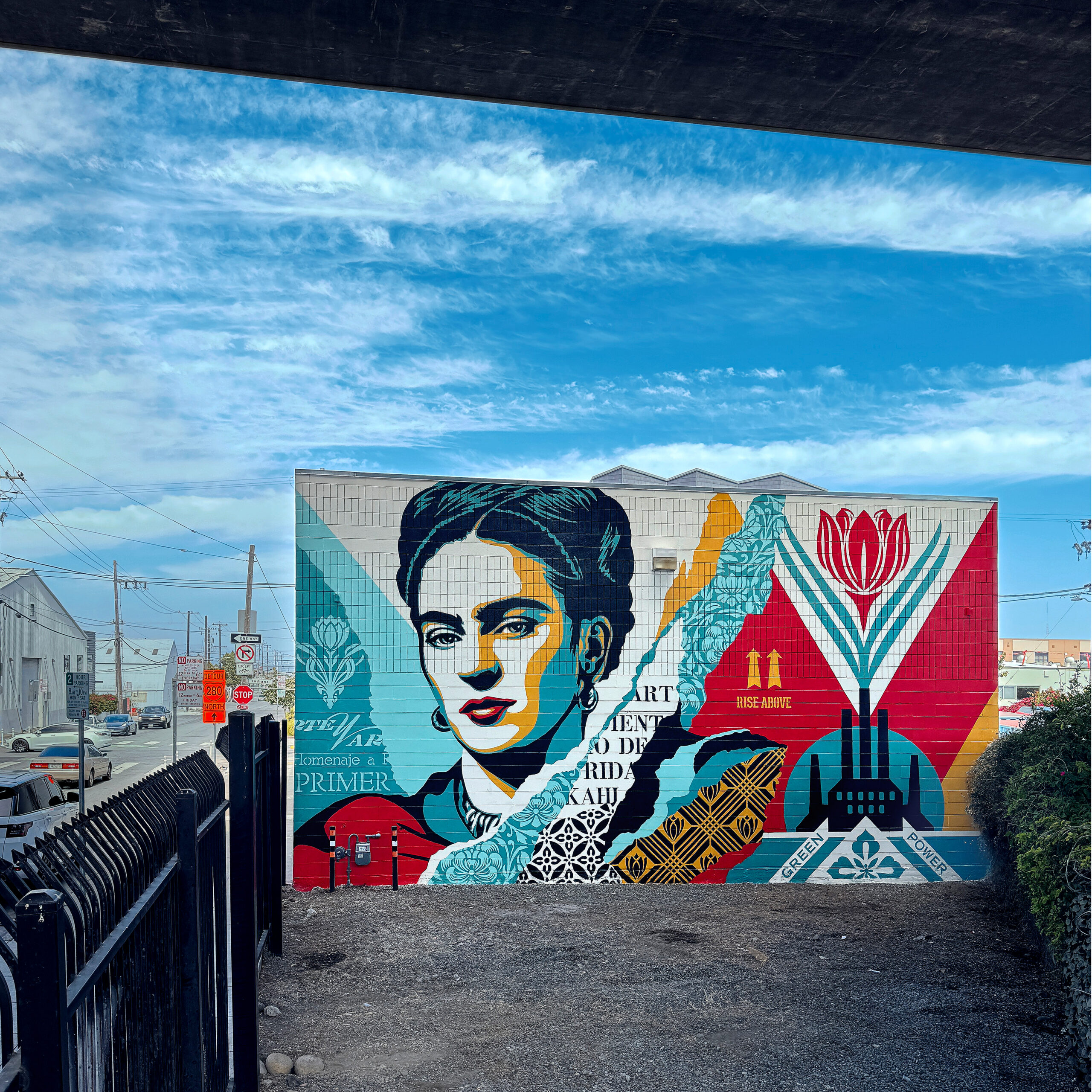 A large mural of Frida Kahlo with colorful geometric patterns on the side of a building under a blue sky.
