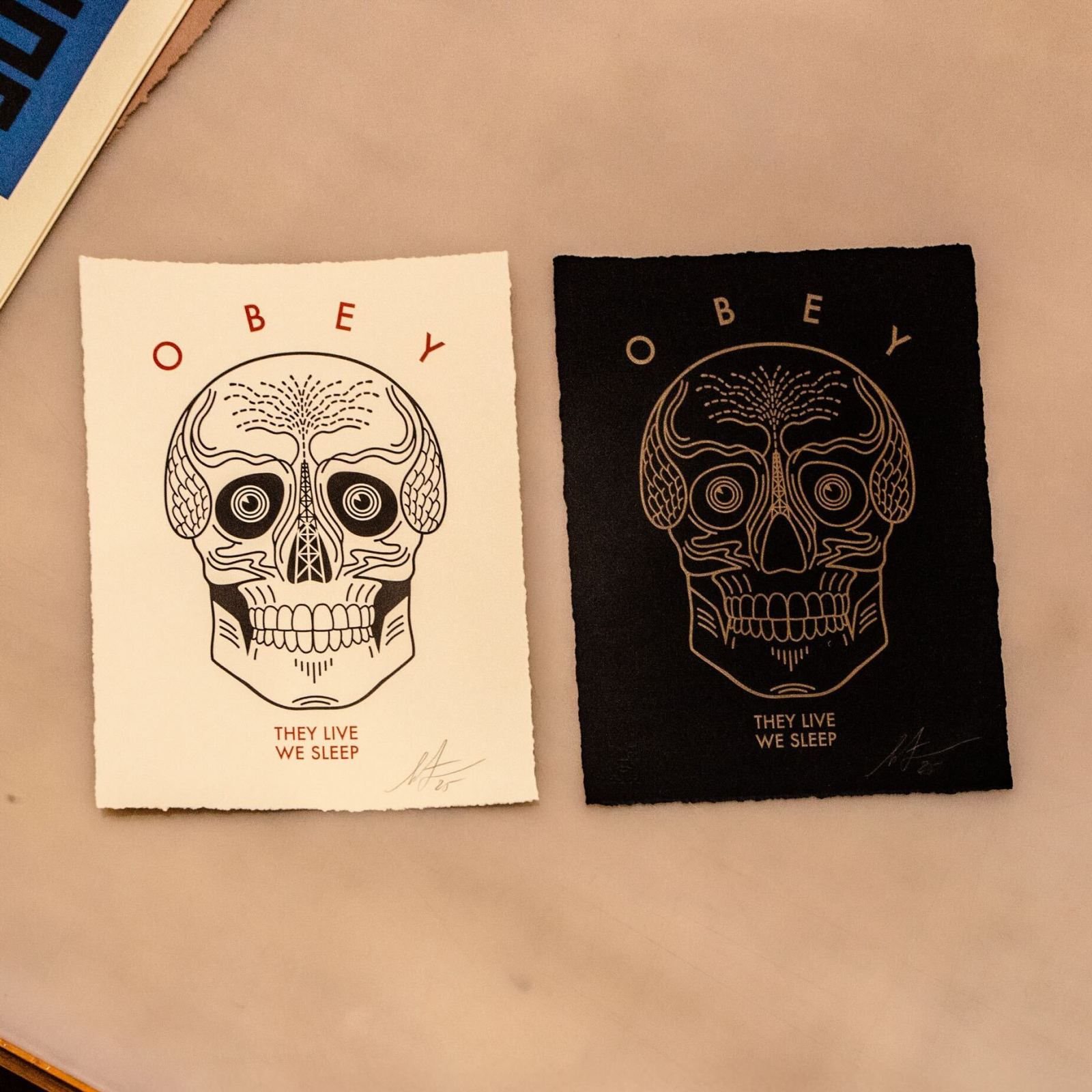 Two OBEY prints: beige & black, each featuring a skull graphic with an oil derrick in its forehead; They Live We Sleep below.