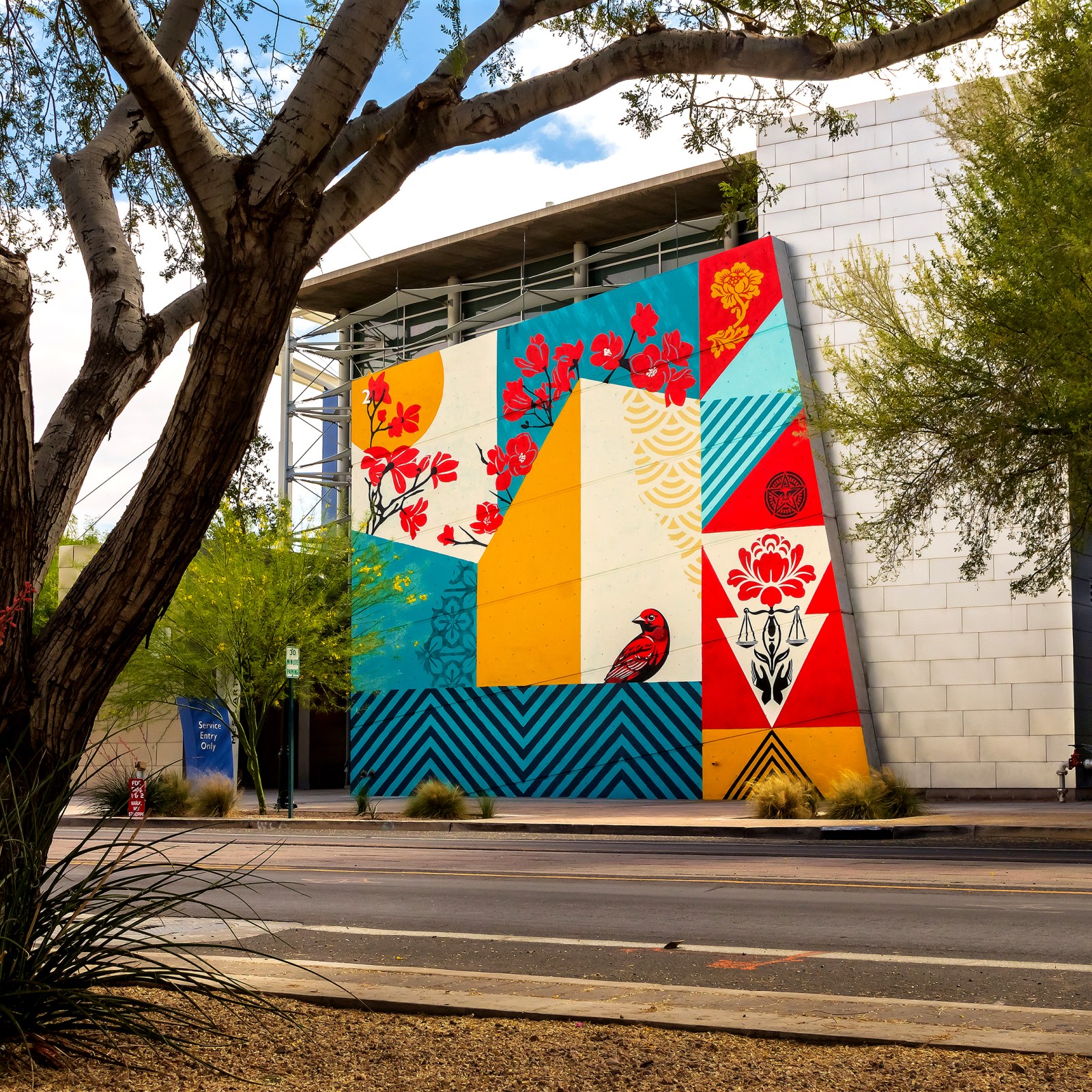 A vibrant mural on a building featuring geometric patterns, red flowers, and a bird, in red, yellow, blue, and cream colors…