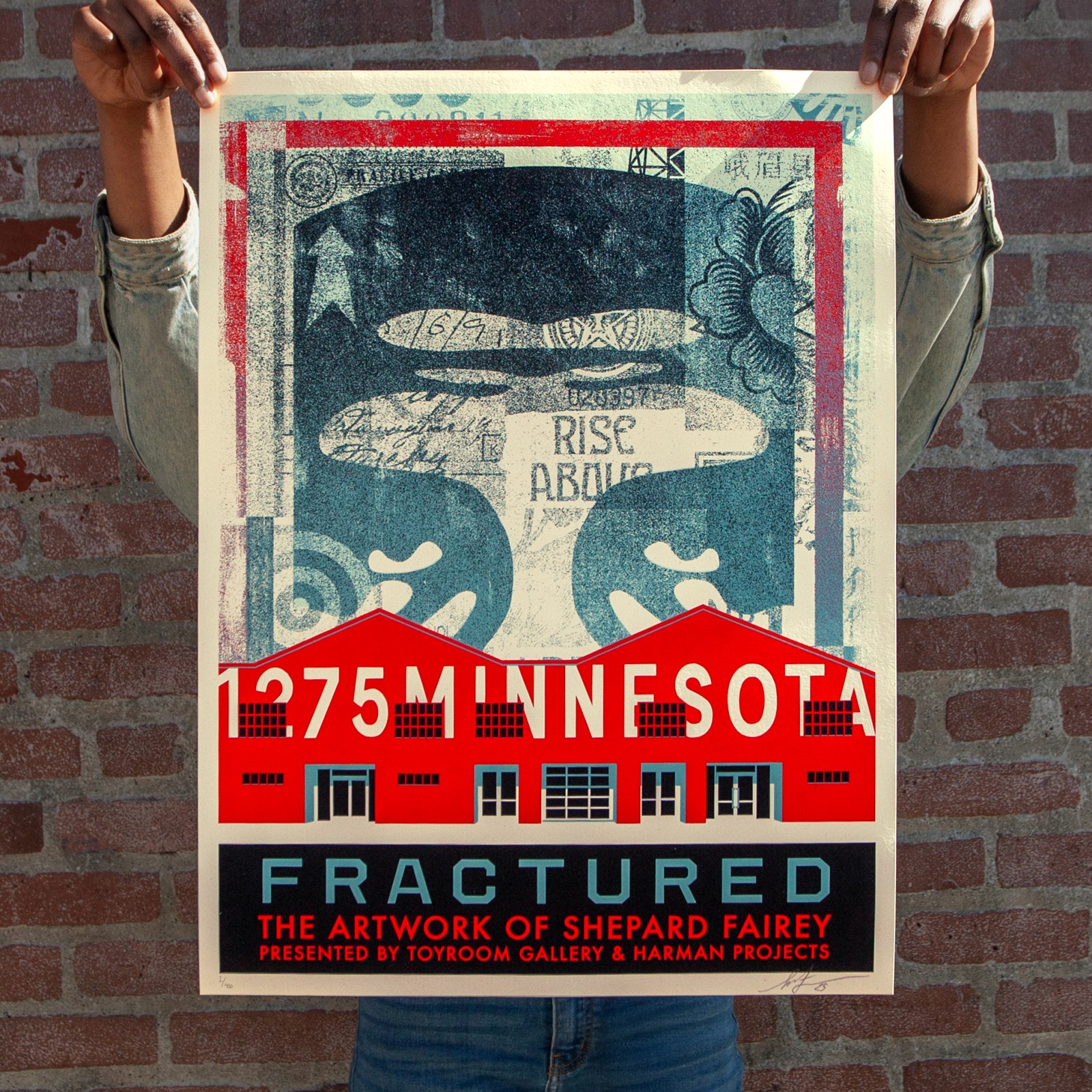 A person holds a poster for 'Fractured: The Artwork of Shepard Fairey.' It features two red buildings, '1275 Minnesota,' and…
