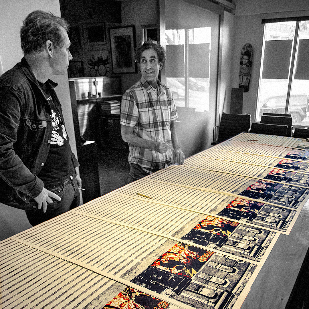 Two men, Shepard Fairey and Glen E. Friedman, standing next to a long table covered with