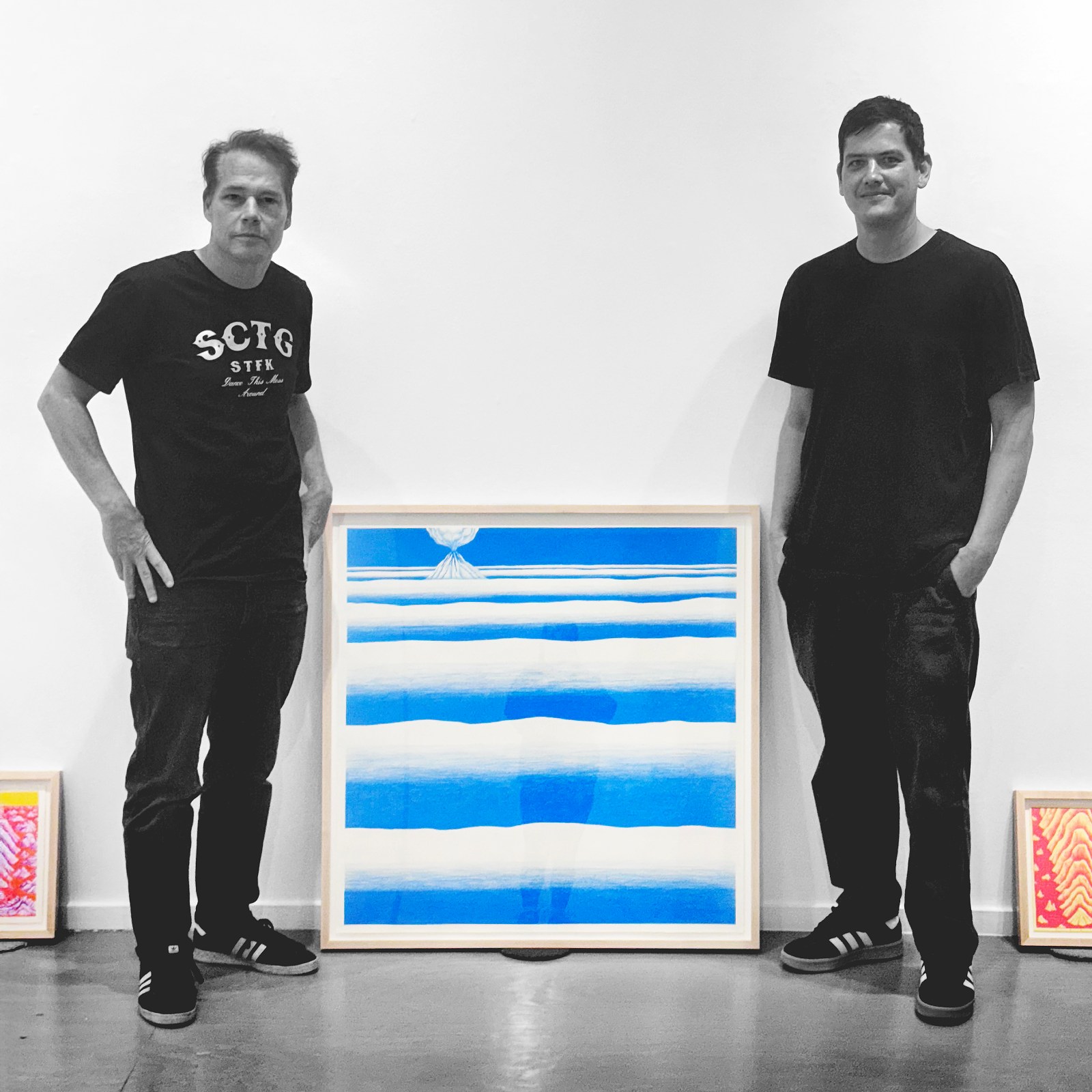 Two men stand on either side of a framed artwork featuring blue and white horizontal stripes with a small white dome at the…