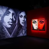 A dimly lit art gallery with large murals of women's faces on the left wall and two illuminated red and black posters on the…