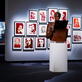 A gallery wall filled with framed pop art portraits in red and white, with a brown bust of a man reading a book on a white…