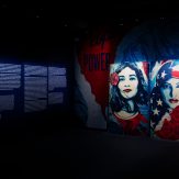 A dimly lit art gallery with a large text panel on the left wall and two illuminated posters of women's faces on the right…