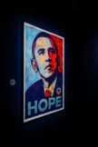 A dimly lit gallery with a single illuminated poster of Barack Obama in red, white, and blue, with the word "HOPE" below him.