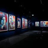 A dimly lit art gallery with a row of illuminated framed portraits of women on the left wall and more artworks in the…