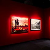 A red-walled art gallery with three illuminated framed artworks: two figures looking at an industrial landscape, a black and…