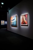 A dimly lit art gallery with two illuminated framed artworks on a white wall: a colorful seascape and a portrait of a woman…