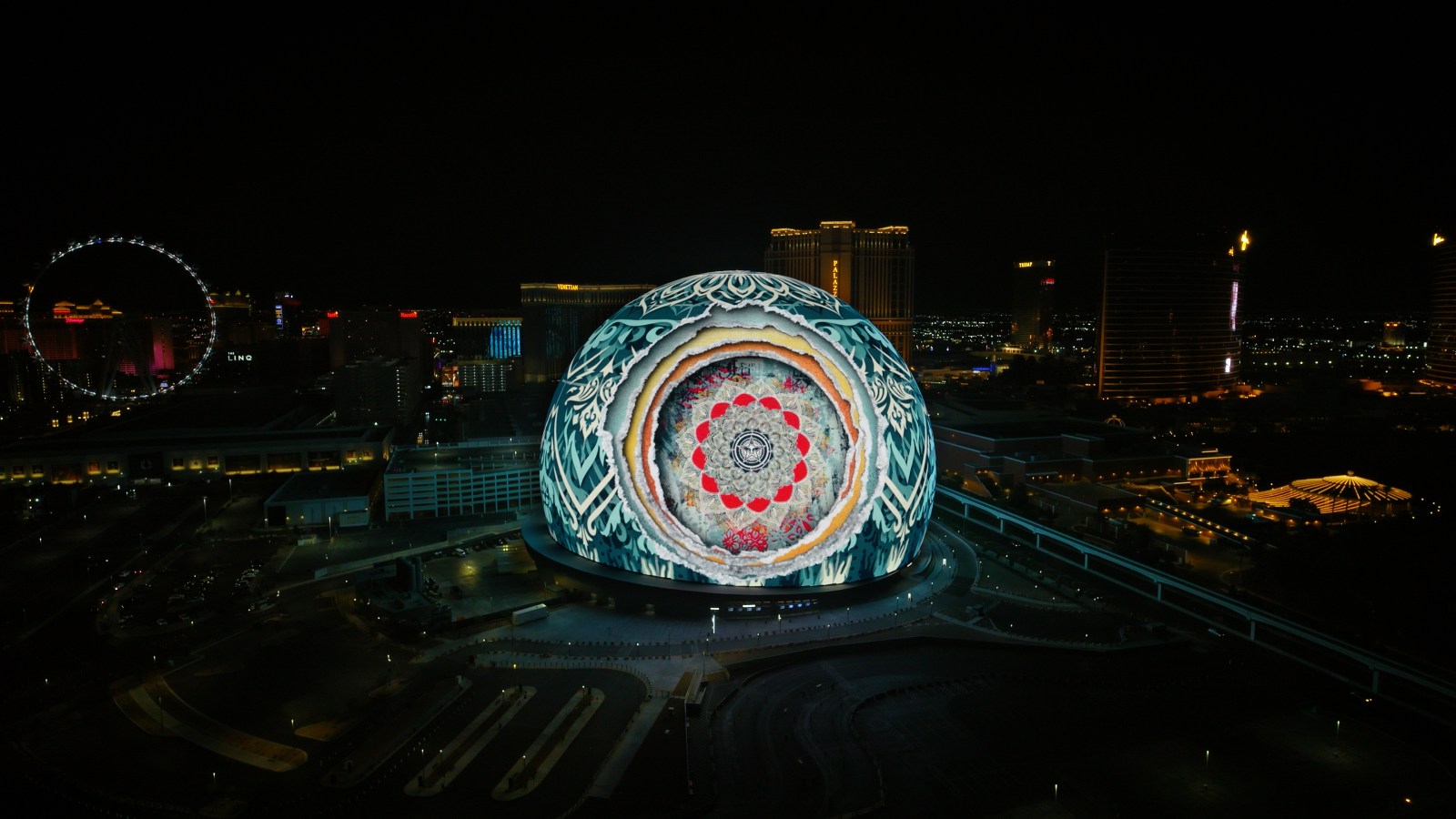An aerial view of a large spherical building illuminated with a colorful, intricate mandala-like projection at night…