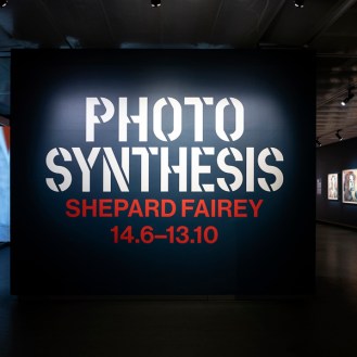 A large black wall with white text "PHOTO SYNTHESIS SHEPARD FAIREY 14.6-13.10" in an art exhibition hall.