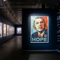 An art exhibition hallway with framed artworks on dark blue walls, including a prominent "HOPE" poster of Barack Obama.
