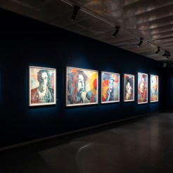 An art exhibition hallway with framed artworks on dark blue walls, including portraits of various figures and a cityscape.