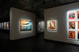 A gallery space with white walls and dark floors. On the left, a framed painting of a sunset over the ocean. On the right, a…