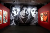 A gallery room with red walls and a large black and white mural featuring three women's faces. A dark bench sits in front of…