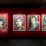 A red gallery wall with four framed art prints. The first reads "MAKE ART NOT WAR" with a woman's face. The others are…