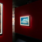 A red gallery wall with a framed artwork of a building in blue and white tones. To the left, a partial view of another…