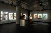 A gallery space with white walls and dark floors, featuring framed art prints and two busts. The busts are of men, one in…