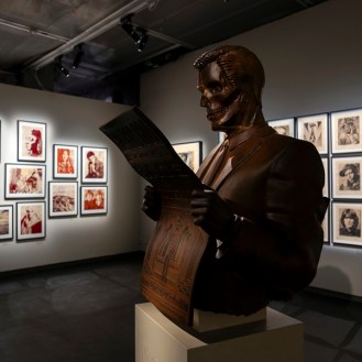 A bronze bust of a man with a skull face, wearing a suit and reading a newspaper, stands in a gallery. Framed art prints…