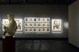 A gallery space with a white bust of a man reading a newspaper in the foreground. Behind it, two large framed artworks and a…