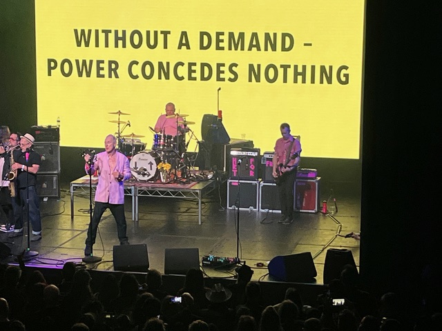 A band performs on stage with a large yellow screen behind them displaying the text: "WITHOUT A DEMAND - POWER CONCEDES…